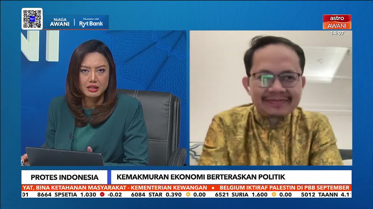 A woman in a green blazer is speaking intently, while a man in glasses and a patterned shirt appears on a split screen. The Astro Awani logo and the time are visible in the corner.
