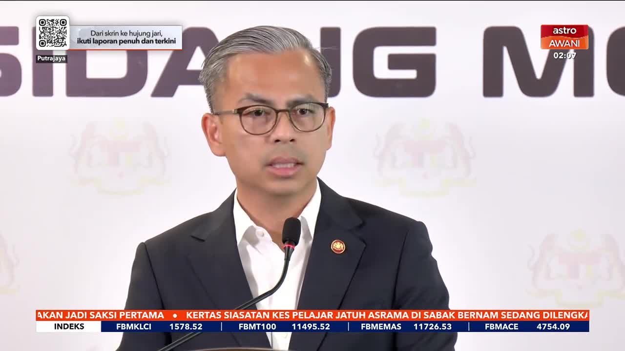 A man in a suit and glasses is speaking into a microphone, the backdrop bearing the words "SIDANG M." The Astro Awani logo is visible in the corner, and financial data scrolls along the bottom of the screen.
