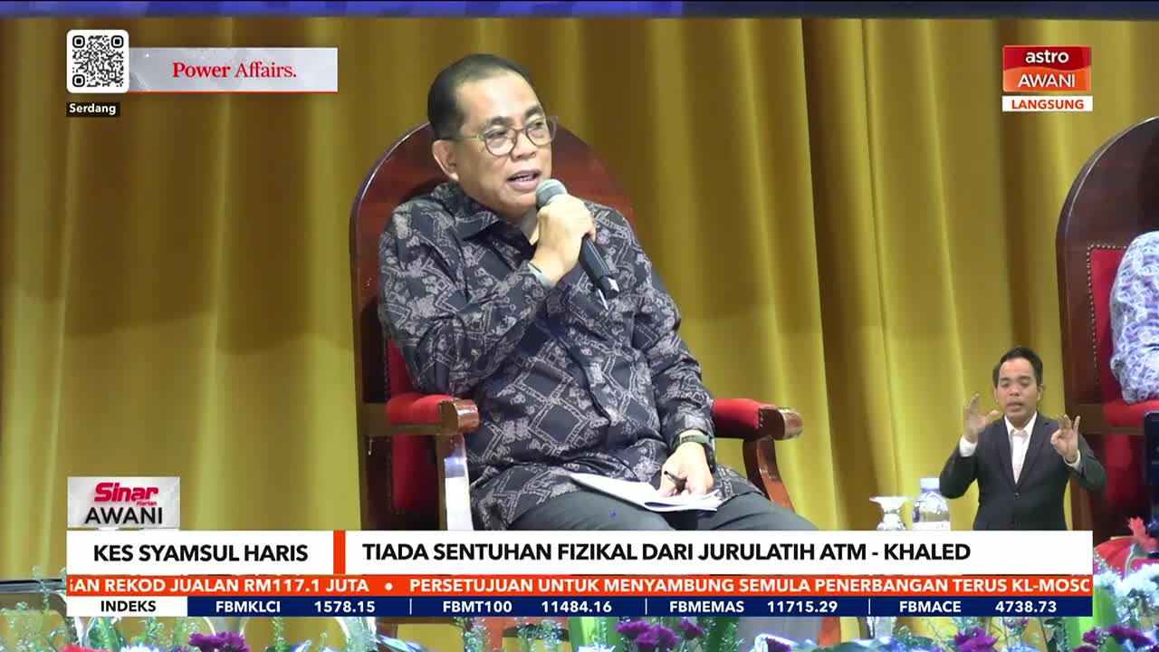 A man in a patterned shirt speaks into a microphone, seated on a large wooden chair. Beside him, another man signs with his hands, appearing on the Astro Awani broadcast from Malaysia.
