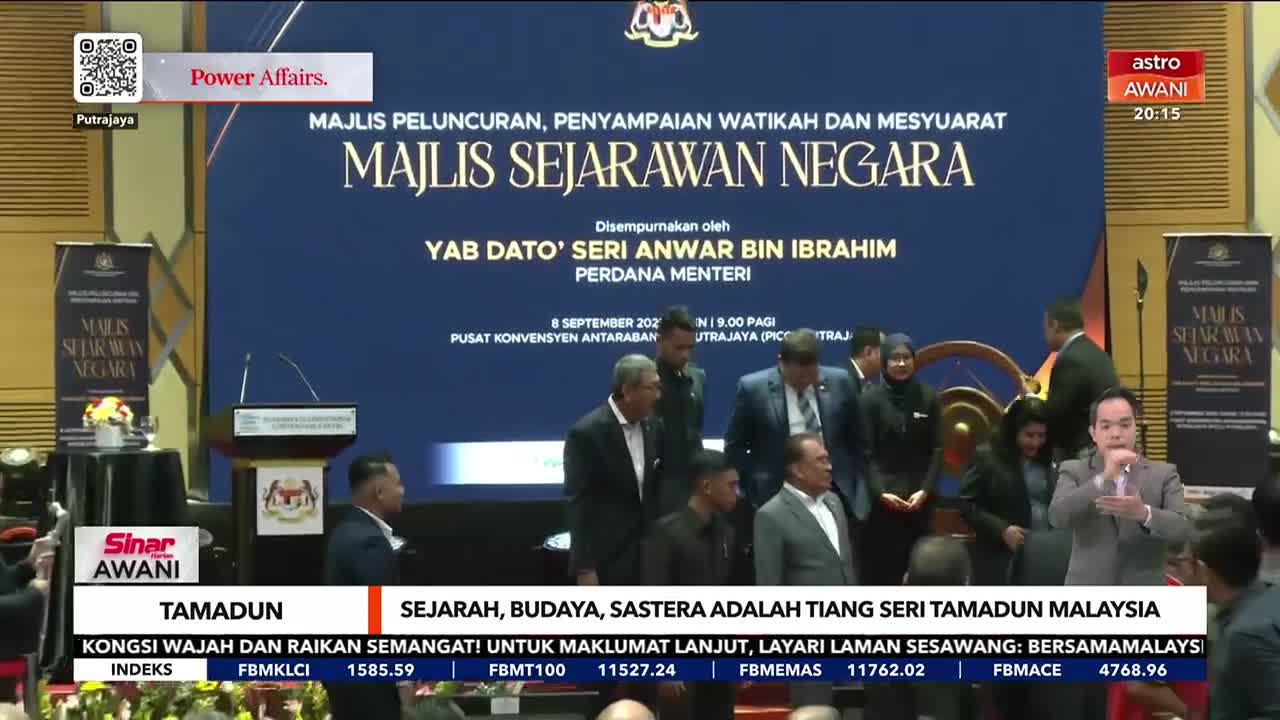 A group of people, including a man in a grey suit, are milling around a stage in Malaysia. A presenter, with a microphone, is speaking in front of the "Astro Awani" logo.
