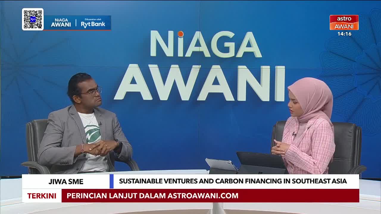 A man in a grey blazer and glasses looks intently toward the woman wearing a pink hijab. The "Niaga Awani" logo and the Astro Awani clock are visible in the studio.

