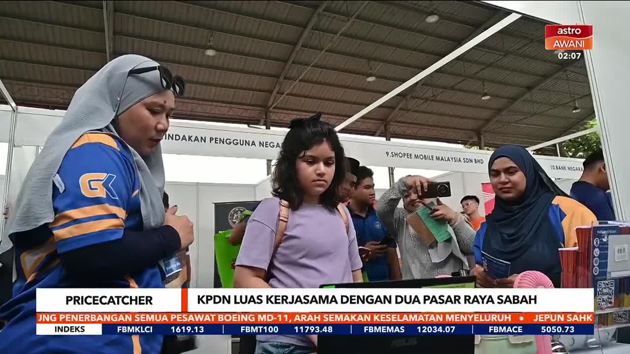 Underneath a large awning, a young girl walks past a booth as a woman in a hijab takes a photo with her phone. The Astro Awani logo is visible in the corner, and a news ticker runs at the bottom of the screen.

