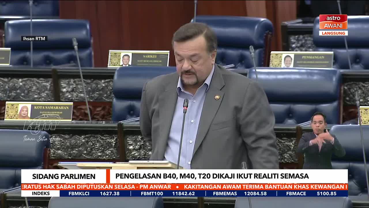 A man in a gray suit speaks into a microphone at a podium, while a sign language interpreter stands to the right. The broadcast is live on Astro Awani from the Malaysian Parliament.
