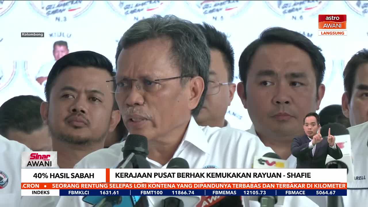A man with glasses speaks into a microphone, surrounded by others, as a Malaysian news broadcast from Astro Awani rolls on. The screen shows a lower third with financial data and a headline about a government appeal.
