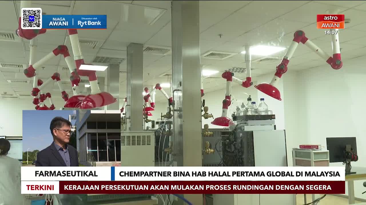 Inside a brightly lit lab, articulated arms with red hoods hang from the ceiling. A news report from Astro Awani in Malaysia is airing, with a man in a suit speaking on a screen within the frame.
