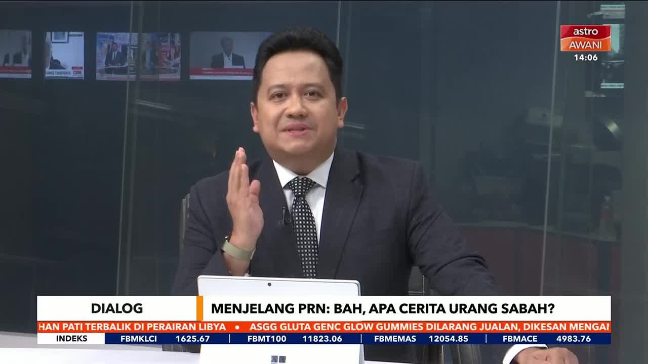 The Astro Awani news anchor, in a suit and tie, gestures with his right hand as he speaks. Below, a ticker displays financial information and news headlines in Malay.

