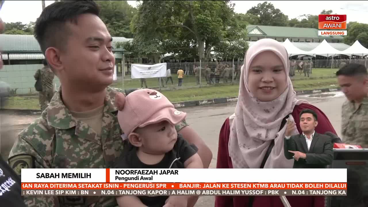 A family, including a baby in a pink hat, is being interviewed on Astro Awani. Behind them, soldiers mill about near a building, likely at a polling location in Sabah.
