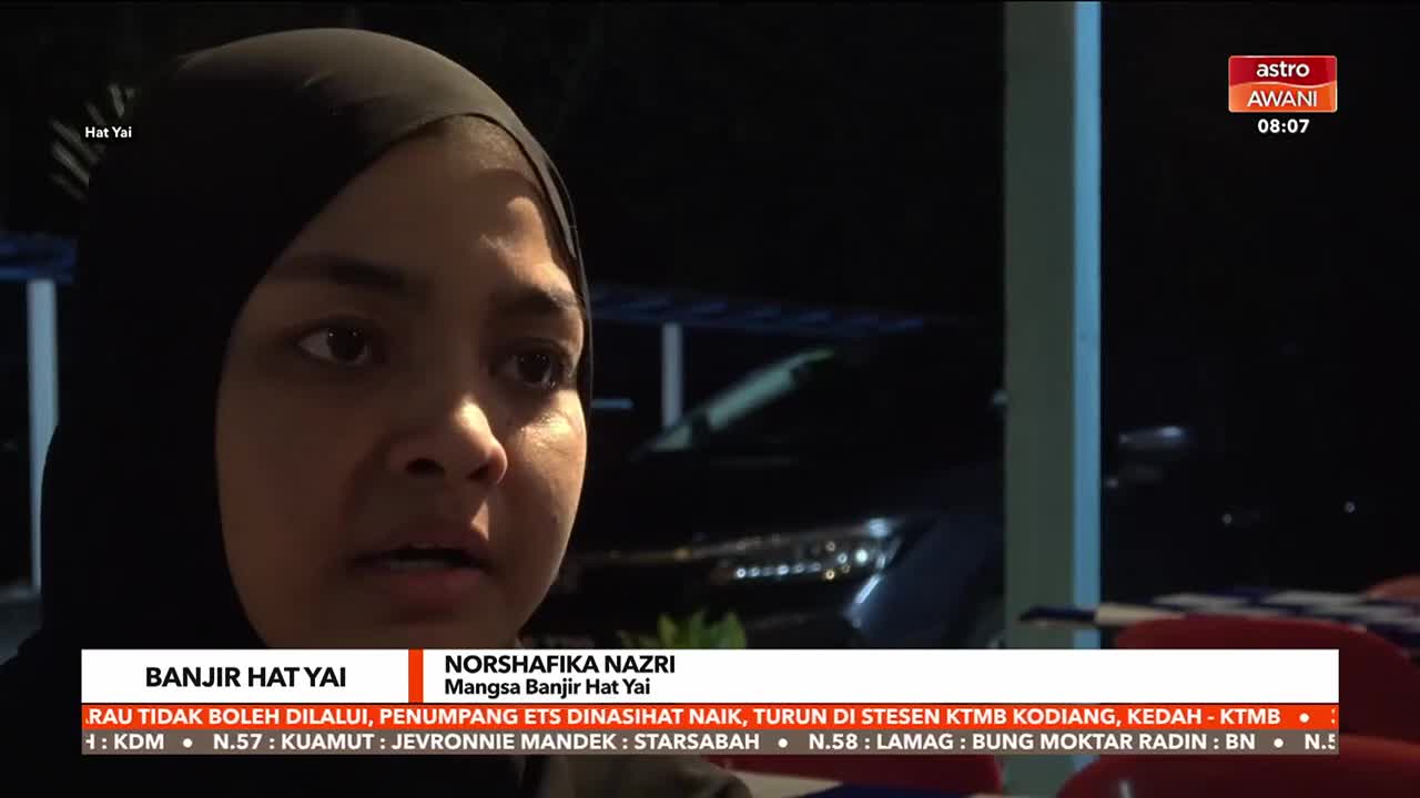 A woman wearing a black hijab speaks earnestly, her face lit by an unseen light. The Astro Awani logo and a news ticker about a flood in Hat Yai are visible on the screen.
