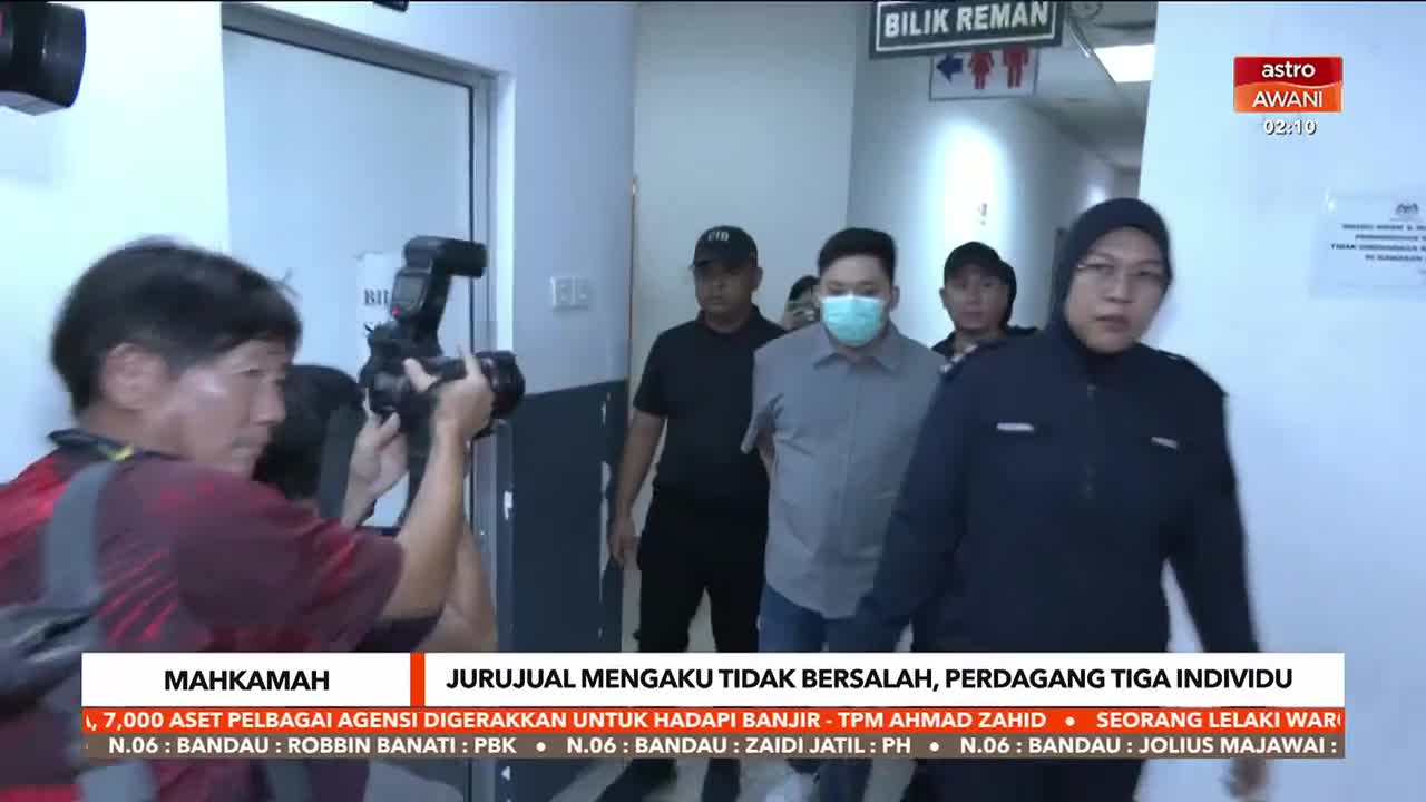 A man in a face mask is escorted down a corridor, flanked by security and followed by a camera crew. The Astro Awani logo is visible, and the headline indicates a court case in Malaysia.
