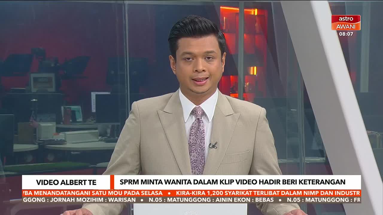 A man in a tan suit and patterned tie speaks directly to the camera, the Astro Awani logo visible in the corner. Below him, text scrolls across a red and black background, offering news headlines.
