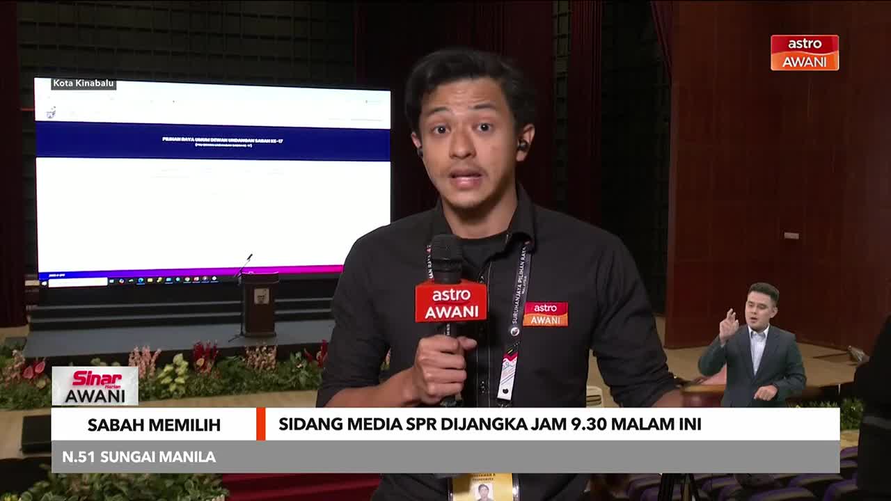 An Astro Awani reporter, microphone in hand, speaks directly to the camera, with a large screen displaying information behind him. To his right, another person in a suit uses sign language.
