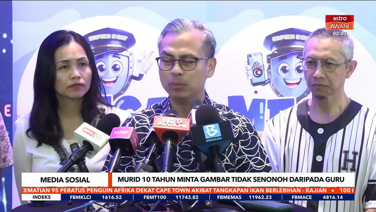 A man in glasses speaks into a cluster of microphones, flanked by two others who listen intently. The Astro Awani logo and a headline about a student and a teacher are visible on the screen behind them.
