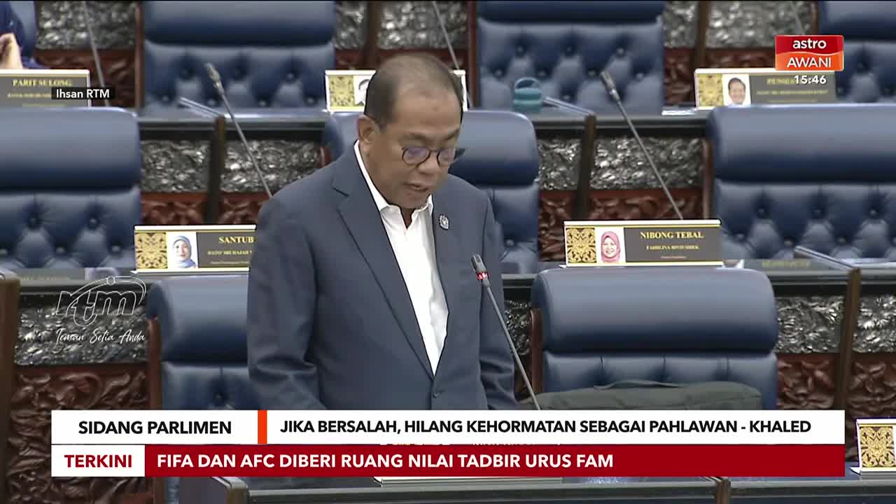 A man in a suit stands at a podium, speaking into a microphone in a room filled with blue upholstered seats. The Astro Awani chyron at the bottom of the screen displays parliamentary proceedings. A man in a suit stands at a podium, speaking into a microphone in a room filled with blue upholstered seats. The Astro Awani chyron at the bottom of the screen displays parliamentary proceedings.