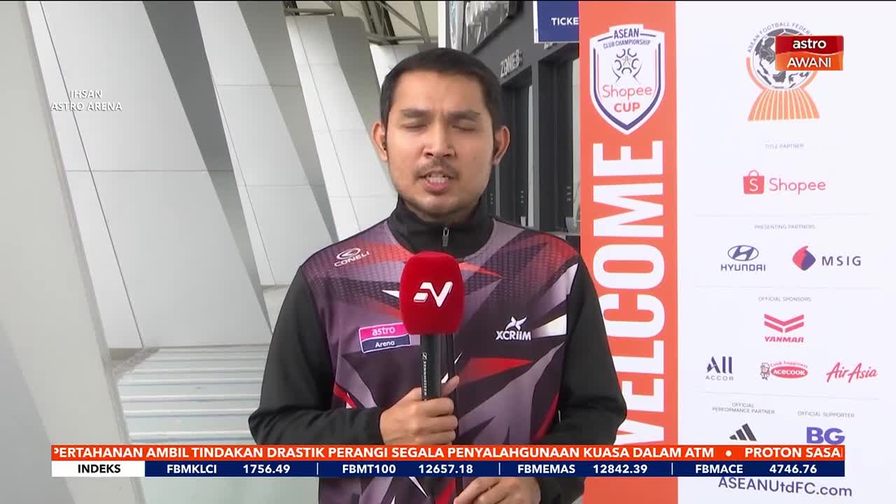 A reporter stands holding a microphone, speaking into it with a large "WELCOME" banner behind him. The banner displays the Shopee Cup logo and several sponsor logos, including Astro Awani. A reporter stands holding a microphone, speaking into it with a large "WELCOME" banner behind him. The banner displays the Shopee Cup logo and several sponsor logos, including Astro Awani.