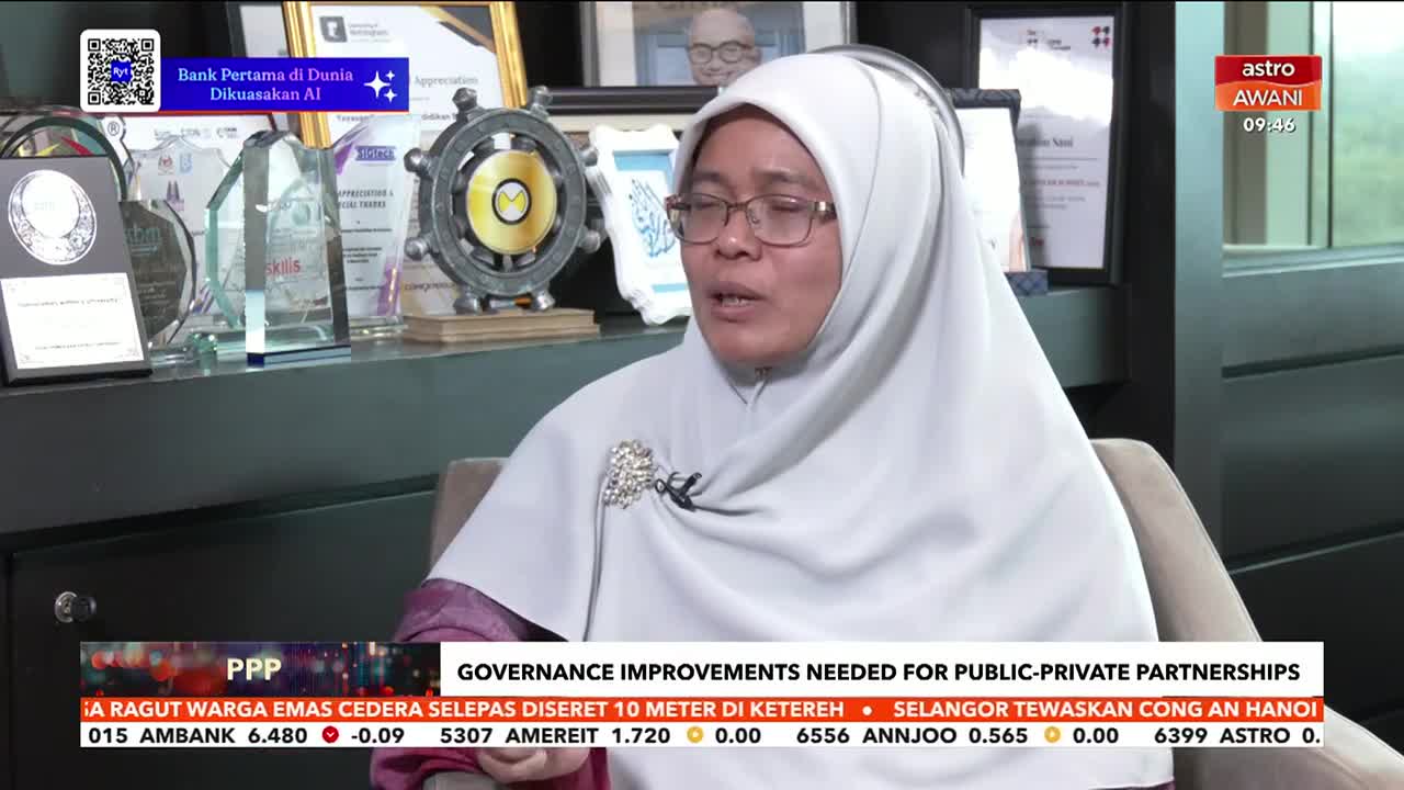 A woman in a light grey hijab speaks, her glasses reflecting the studio lights. Behind her, an array of awards gleams on a shelf, and a ticker displaying financial news scrolls across the bottom of the screen, indicating this is a segment on Astro Awani. A woman in a light grey hijab speaks, her glasses reflecting the studio lights. Behind her, an array of awards gleams on a shelf, and a ticker displaying financial news scrolls across the bottom of the screen, indicating this is a segment on Astro Awani.