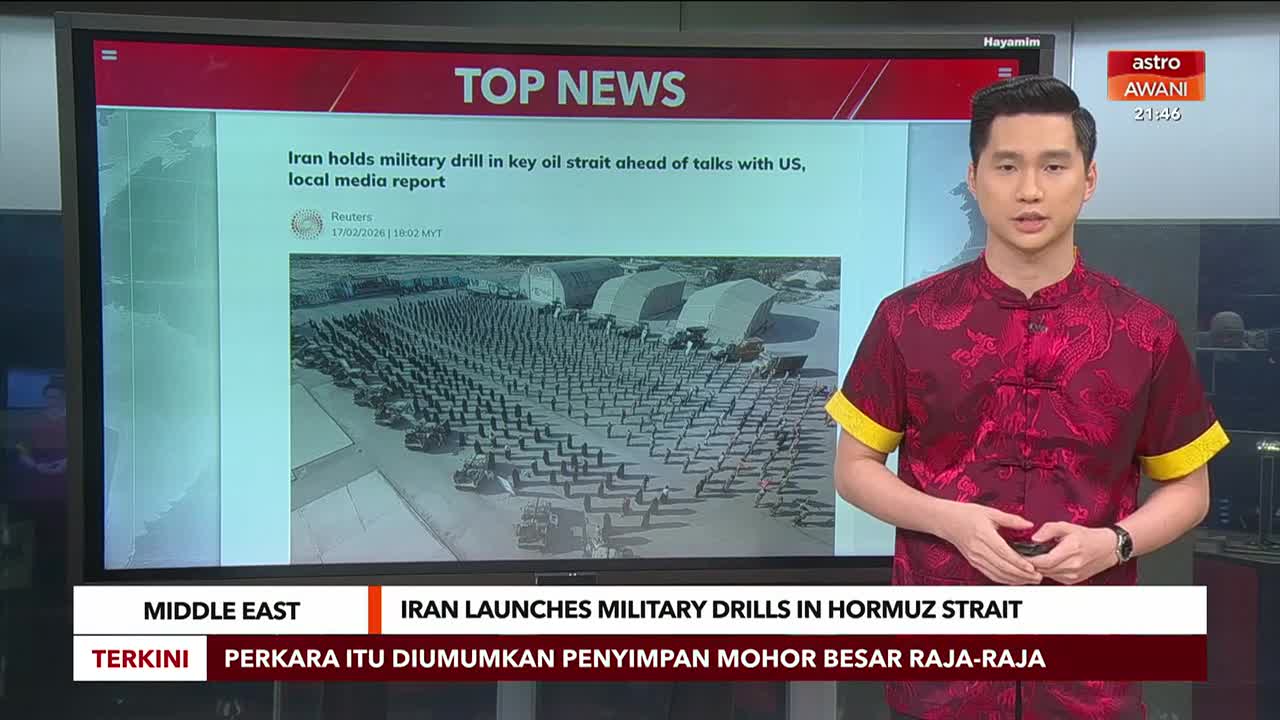 A man in a red patterned shirt stands next to a large screen displaying a news report from Astro Awani about Iran's military drills. The screen shows an aerial view of numerous soldiers and vehicles lined up in formation. A man in a red patterned shirt stands next to a large screen displaying a news report from Astro Awani about Iran's military drills. The screen shows an aerial view of numerous soldiers and vehicles lined up in formation.