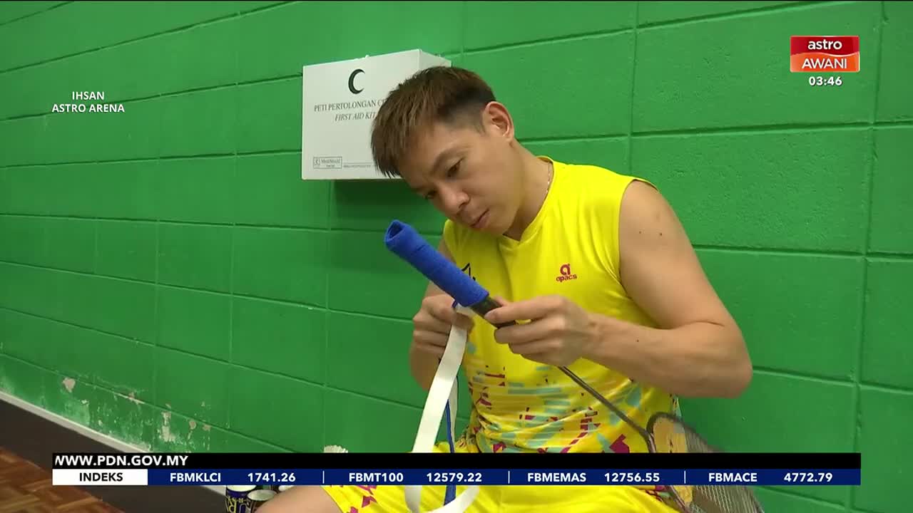 The badminton player carefully wraps white tape around the blue grip of his racket. On the screen, Astro Awani reports the time as 03:46. The badminton player carefully wraps white tape around the blue grip of his racket. On the screen, Astro Awani reports the time as 03:46.