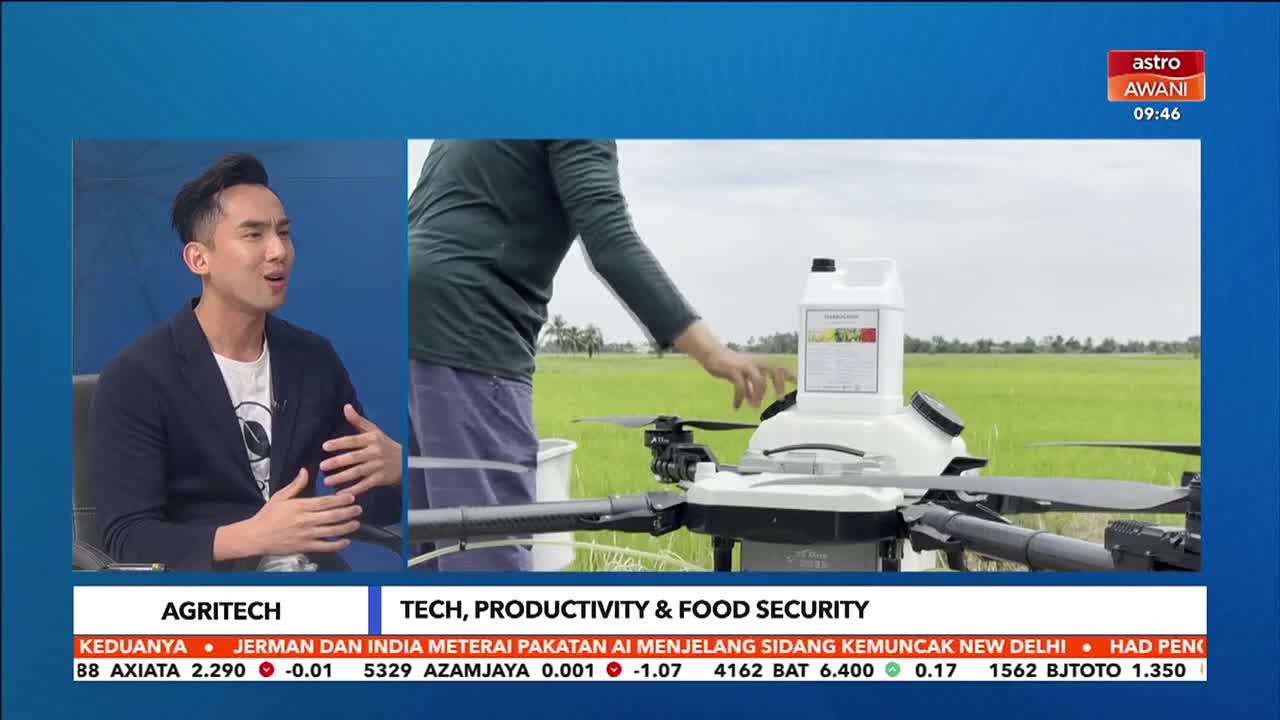 A person in a dark shirt and blue pants is adjusting a large drone in a green field, with a white tank mounted on top. The Astro Awani broadcast shows this scene alongside a man in a suit discussing agritech. A person in a dark shirt and blue pants is adjusting a large drone in a green field, with a white tank mounted on top. The Astro Awani broadcast shows this scene alongside a man in a suit discussing agritech.