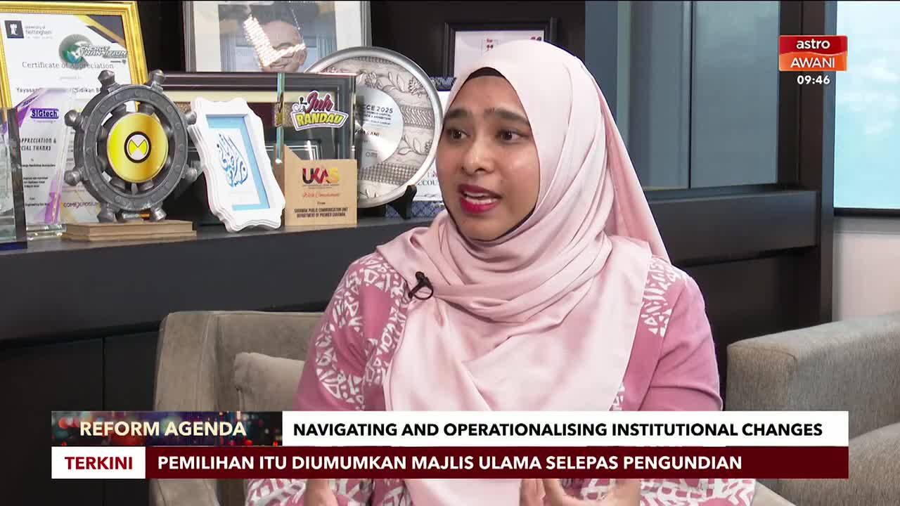 A woman in a pink hijab speaks animatedly, her gaze directed slightly off-camera. Behind her, a shelf displays various awards and a framed piece of calligraphy, all under the Astro Awani logo.