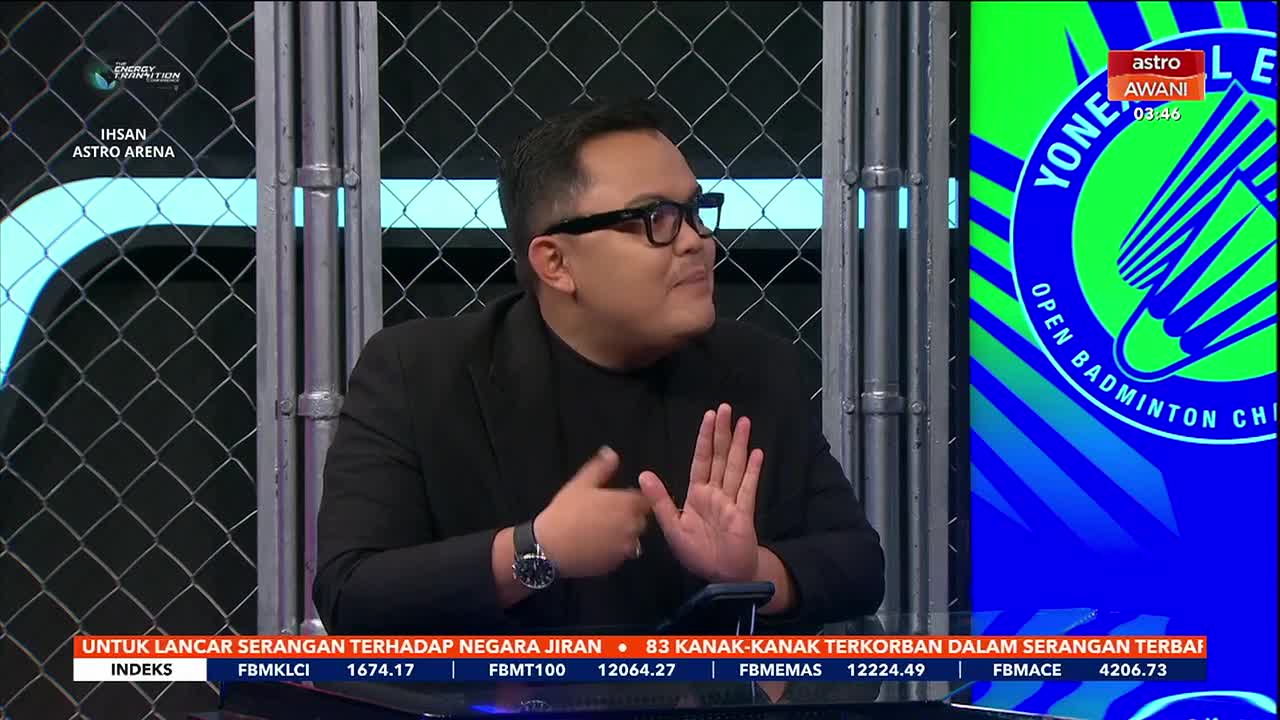 A man in a black suit and glasses gestures with his hands while speaking. Behind him, a screen displays the Astro Awani logo and information about the Yonex Open Badminton Championship.