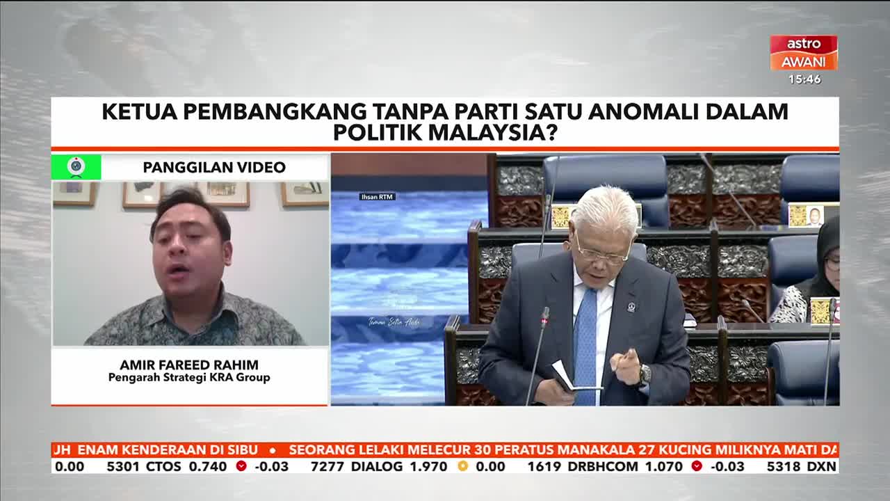 A man in a suit speaks animatedly in a parliamentary setting, while another man appears on a video call. The Astro Awani chyron asks if a party-less development chief is an anomaly in Malaysian politics.