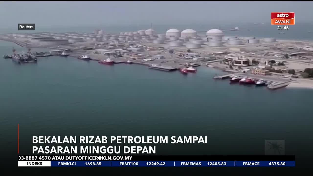 A line of red and white boats are docked along the shore of a large industrial port. Beyond them, rows of white storage tanks stretch across the hazy horizon.