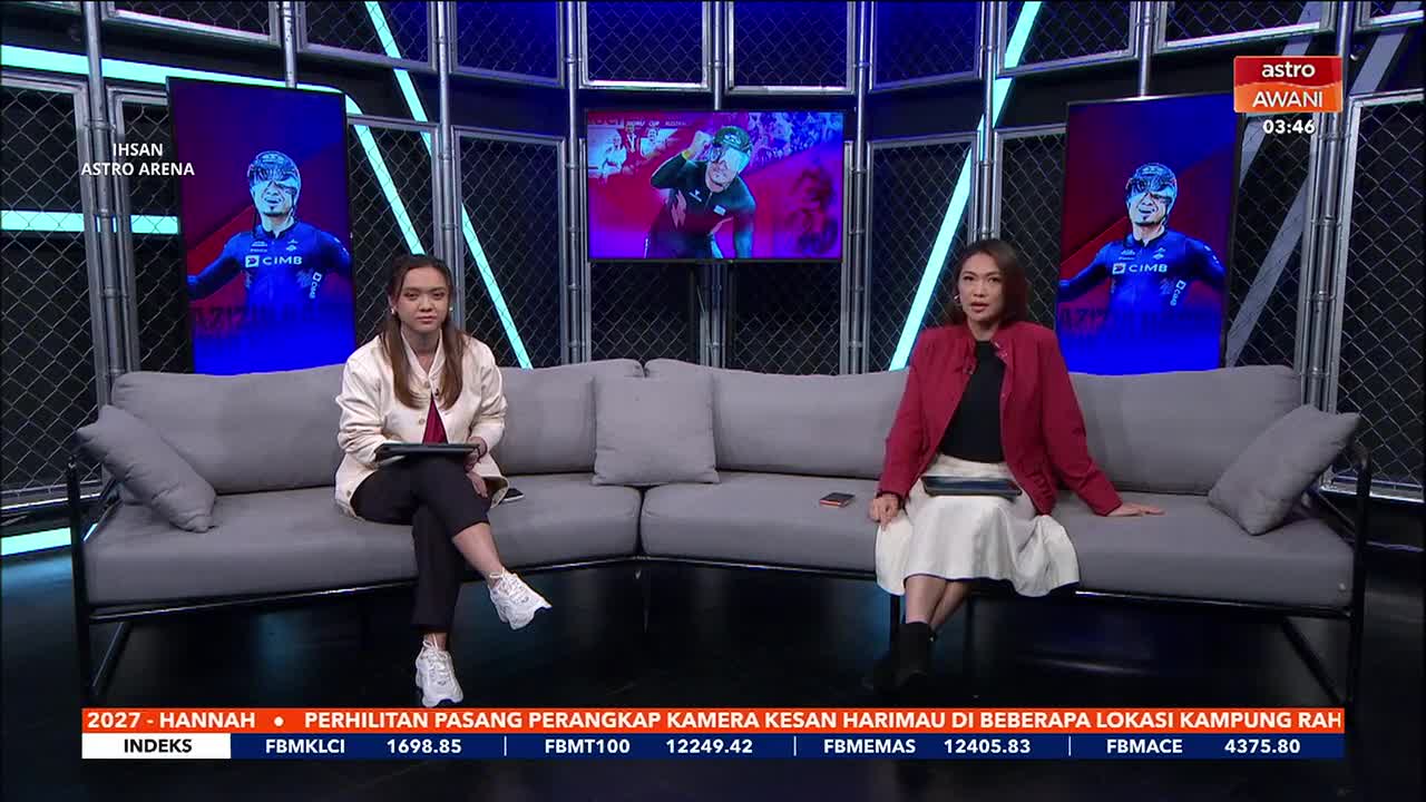 Two women sit on a grey couch in a studio, each holding a tablet. Behind them, a large screen displays a man in a cycling outfit, and another screen shows a close-up of the same cyclist. A ticker runs along the bottom of the screen with Malaysian stock market data.