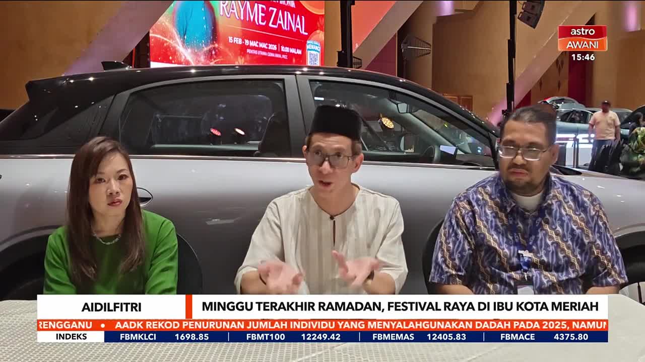 A man in a songkok gestures as he speaks, while a woman in green and another man in a patterned shirt listen. Behind them, a silver car gleams under bright lights, with a large screen displaying "RAYME ZAINAL" in the background.