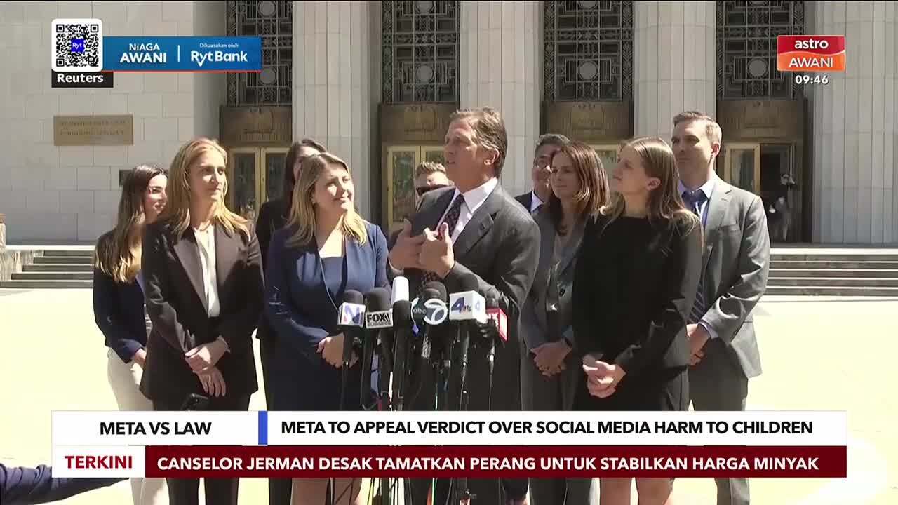 A man in a suit speaks animatedly into a cluster of microphones, while a group of people stand behind him.  The Astro Awani news ticker scrolls across the bottom of the screen.
