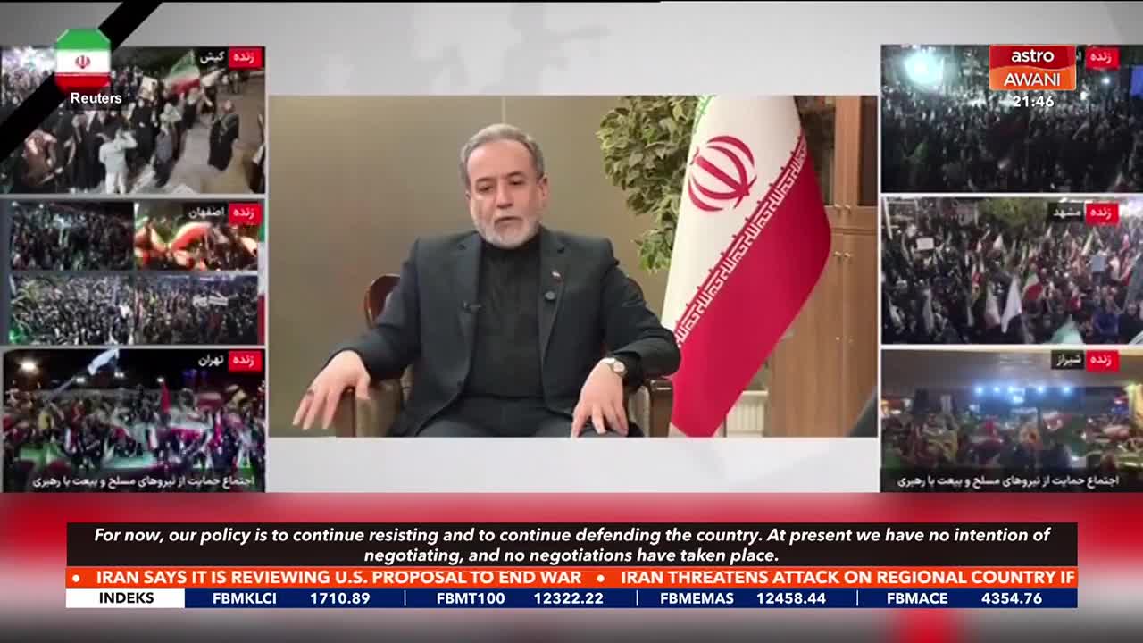 A man in a suit sits in a chair, speaking. On the right, crowds of people wave flags under bright lights, with "astro AWANI" visible.