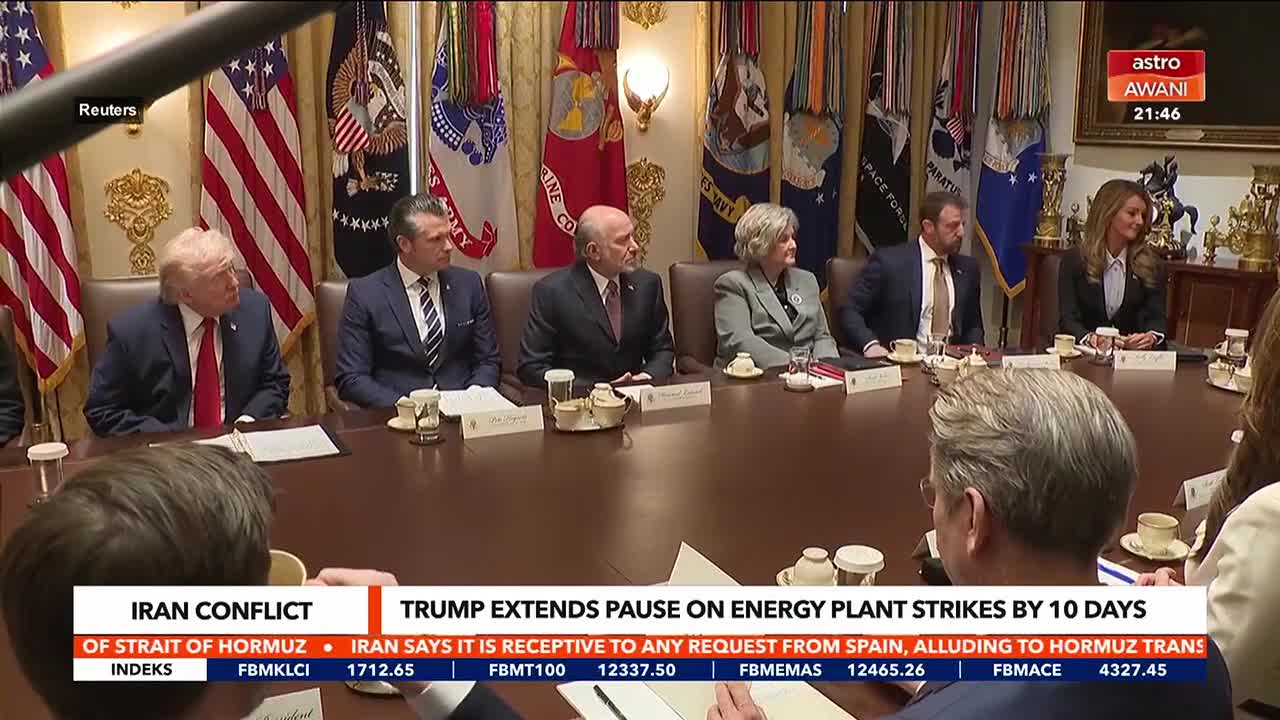A group of people in suits sit around a large table, their attention directed towards the left. Behind them, American flags and military service flags line the wall. A news ticker at the bottom of the screen, with the Astro Awani logo, displays headlines about the Iran conflict.