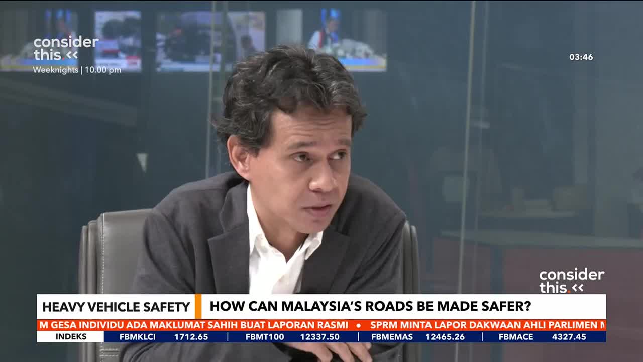 A man in a dark suit over a white shirt speaks, his brow furrowed in thought. Below him, a chyron displays "HEAVY VEHICLE SAFETY" and "HOW CAN MALAYSIA'S ROADS BE MADE SAFER?"