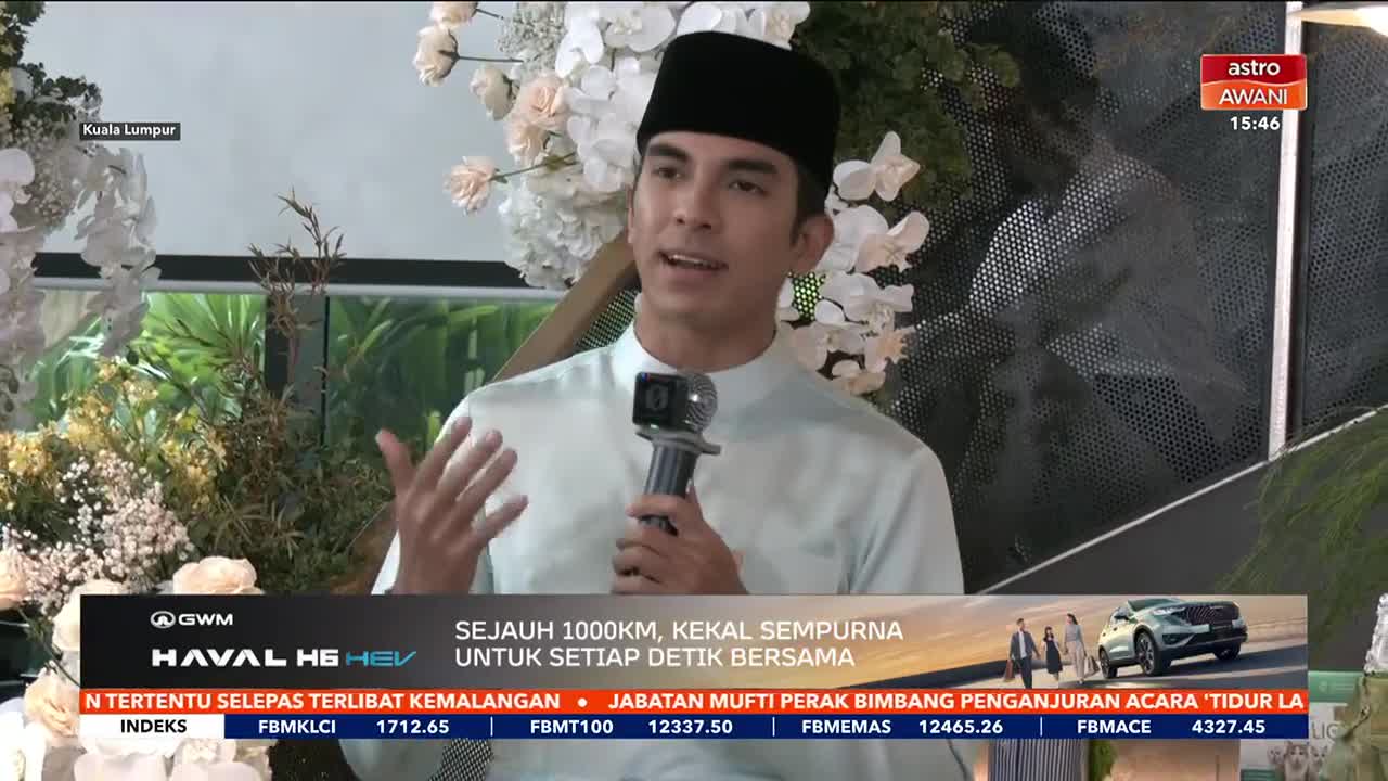 A man in a light blue shirt and black peci speaks into a microphone, gesturing with his hands. Behind him, a floral arrangement of white and blush pink flowers stands against a backdrop of greenery.