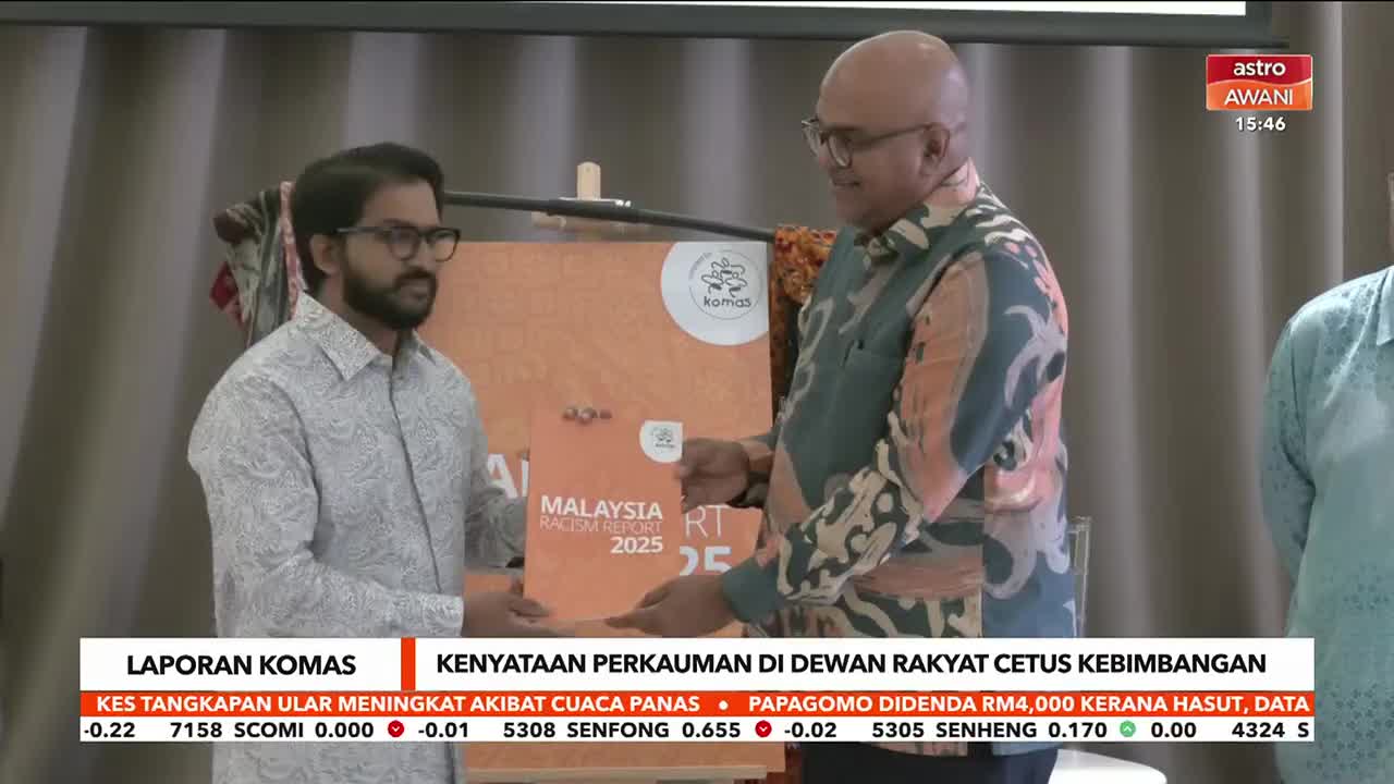 A man in a patterned shirt hands a document to another man in a batik shirt. The Astro Awani logo is visible in the upper right corner.