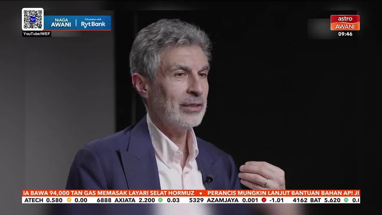 A man with salt-and-pepper hair and a beard speaks with his hands, gesturing as he talks. A ticker tape scrolls across the bottom of the screen, displaying financial news in Malay.
