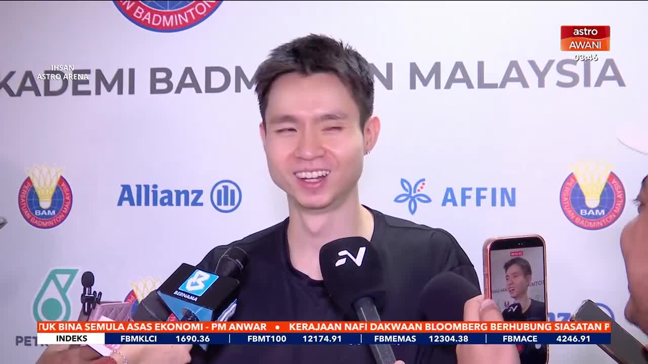 A young man, likely a badminton player, is speaking into microphones held by reporters from Astro Awani. He's smiling broadly, his eyes crinkled, as he answers their questions.