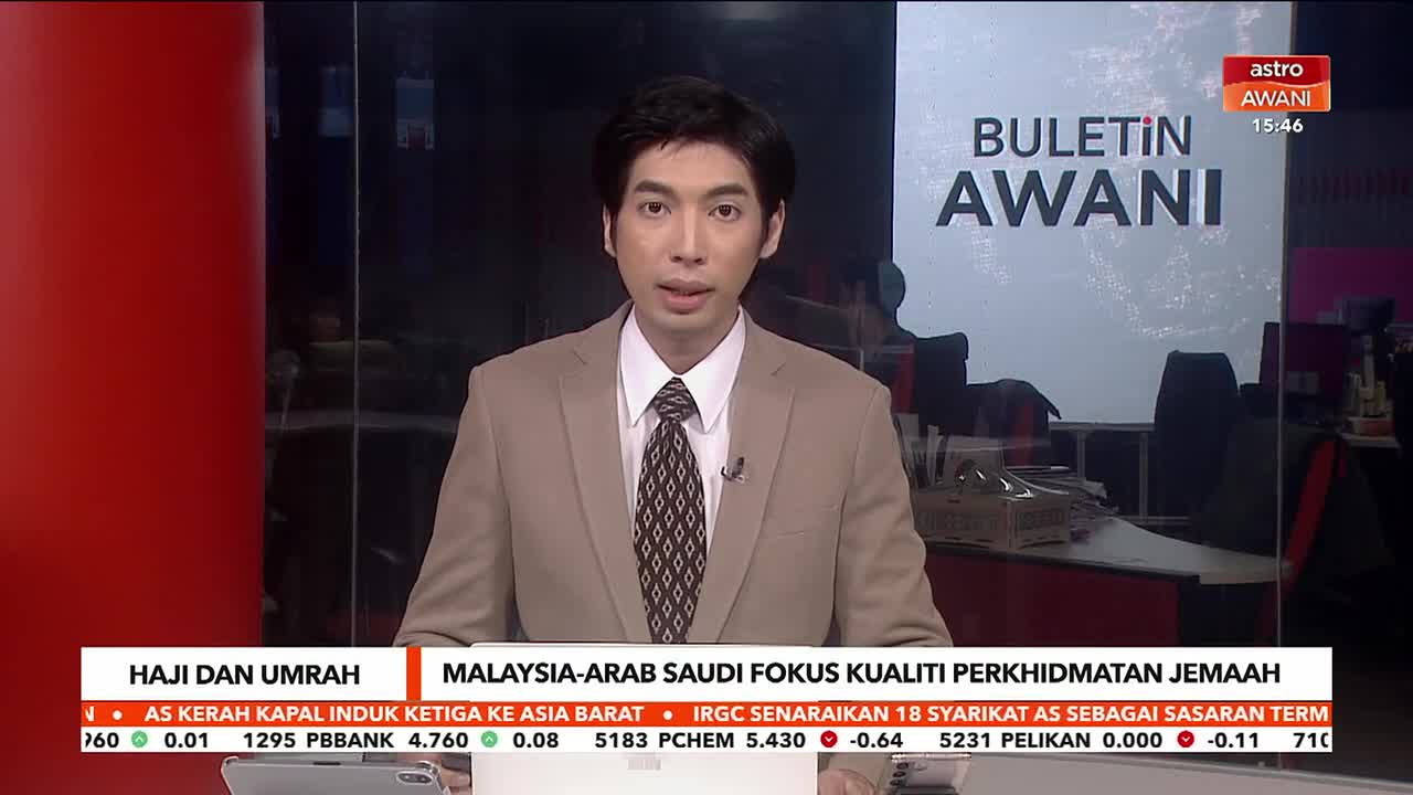 A man in a tan suit stands at a news desk, speaking into a microphone. Behind him, a screen displays "BULETIN AWANI" and a ticker scrolls with financial information.