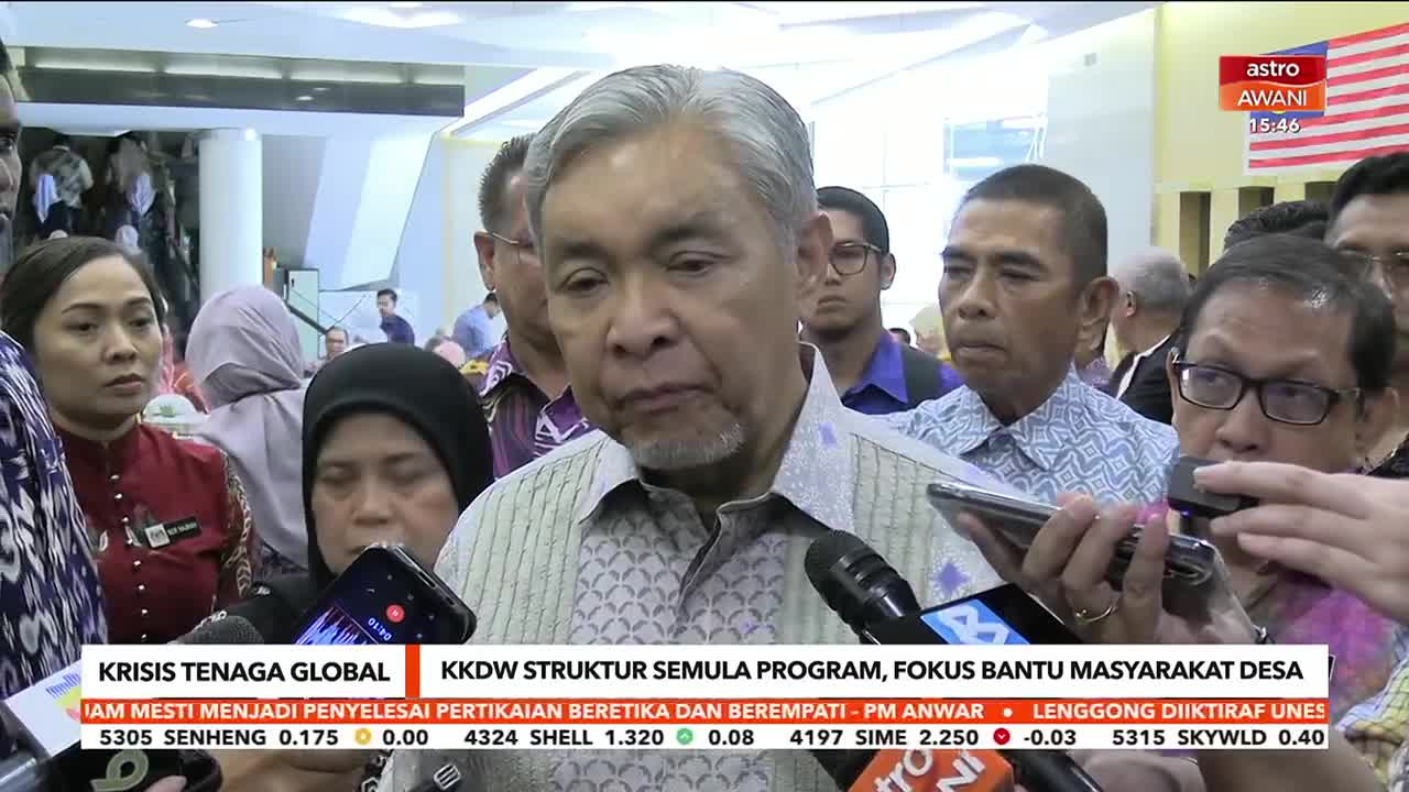 A man in a patterned shirt speaks into a microphone, surrounded by a crowd of people. Microphones from Astro Awani are held close, capturing his words.