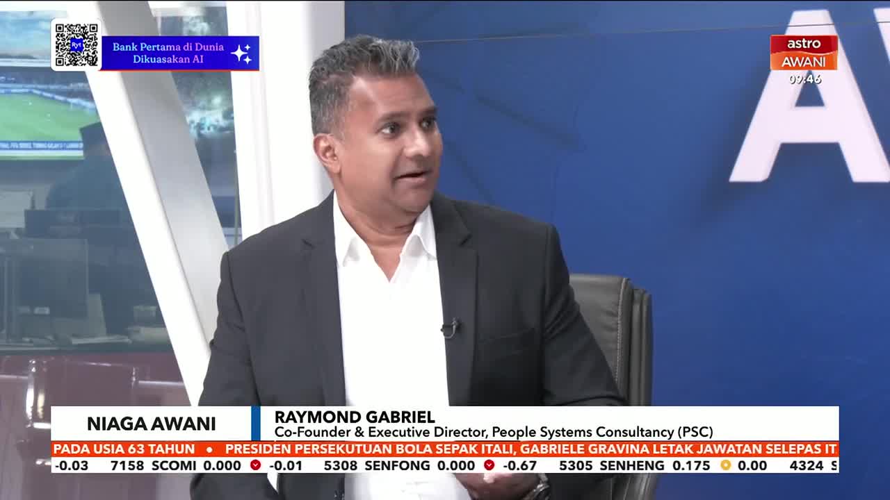 A man in a dark suit and white shirt speaks animatedly during a broadcast on Astro Awani. Below him, a ticker displays financial data and news headlines.