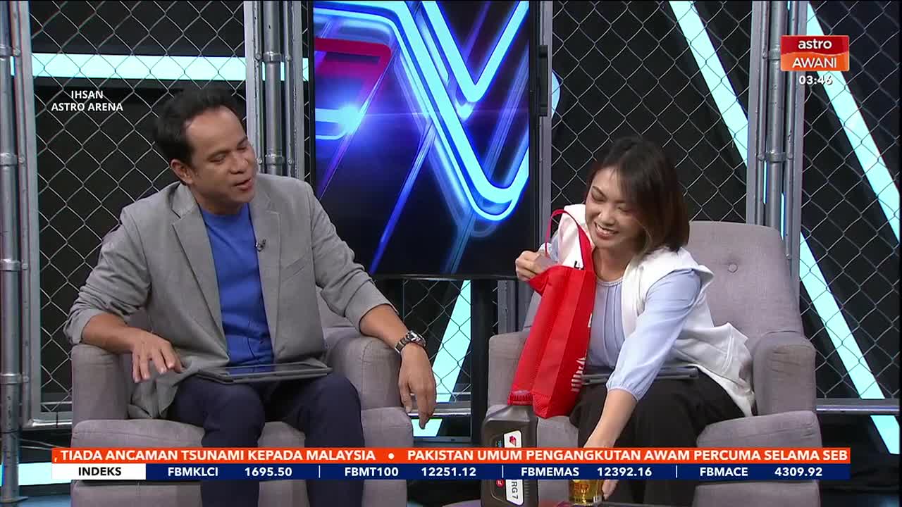 A woman in a white vest pulls a red bag from her lap, her smile wide. Beside her, a man in a grey blazer gestures with his left hand while looking towards her.