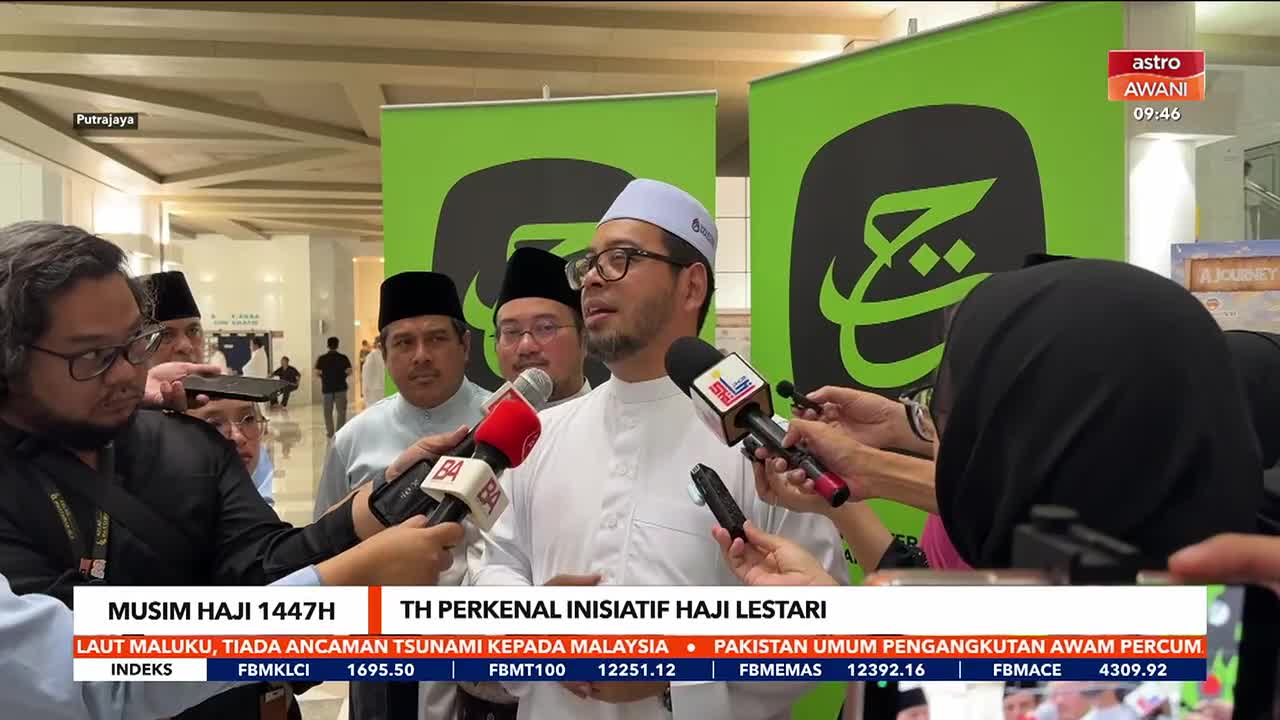 A man in a white tunic and cap speaks into multiple microphones, surrounded by reporters. Behind him, a large green banner displays Arabic calligraphy.