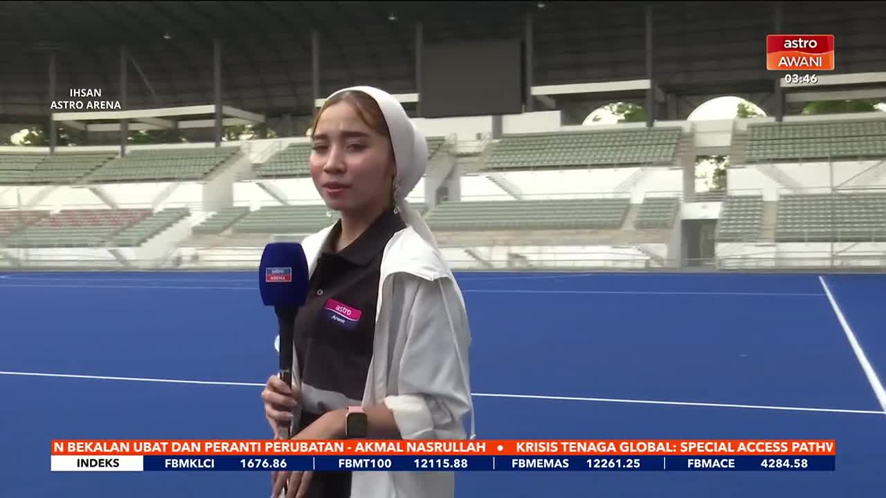A young woman stands on a bright blue field, holding a microphone for Astro Awani. Behind her, empty stadium seats stretch up towards a gray roof.