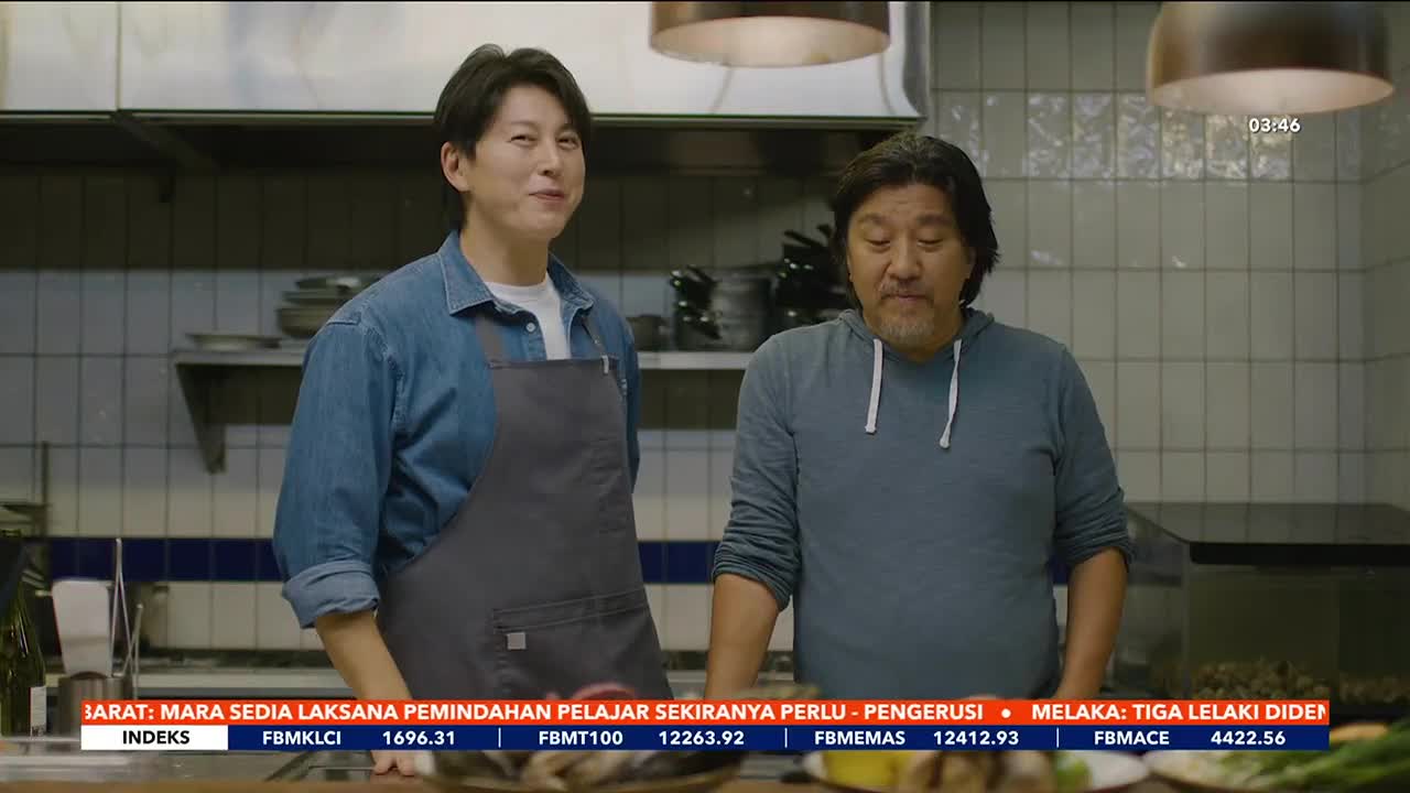 Two men stand in a kitchen, one in a denim shirt and apron, the other in a blue hoodie. A ticker tape with Malaysian financial data scrolls across the bottom of the screen.