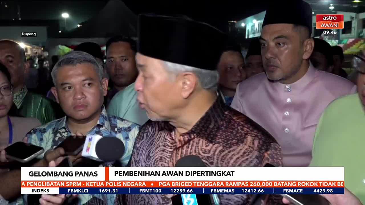 A man in a patterned shirt speaks into a microphone, surrounded by reporters. Another man in a pink Baju Melayu stands nearby, his gaze fixed ahead.