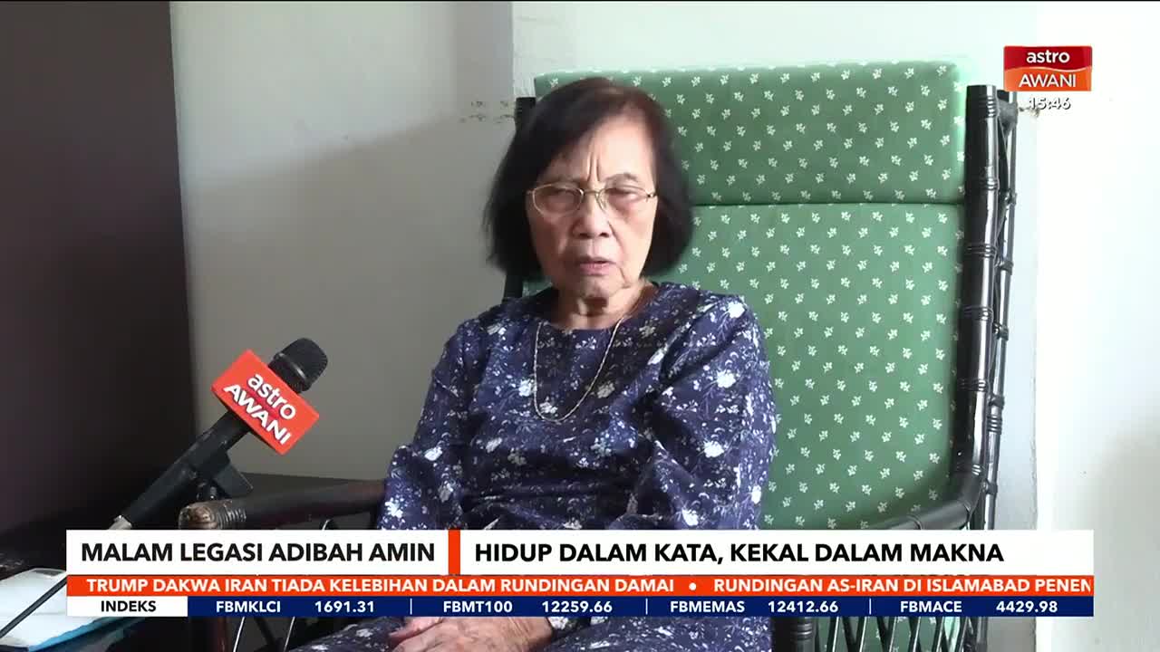 An Astro Awani microphone is positioned in front of an older woman seated in a patterned armchair. The screen displays a news ticker with headlines in Malay.