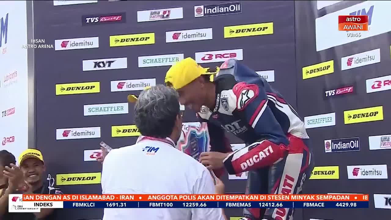 A racer in a red and white suit, wearing a yellow cap, leans in to embrace a man in a white shirt. The background is a wall covered in sponsor logos, including Dunlop and Idemitsu, typical of a Malaysian motorsports event broadcast on Astro Awani.