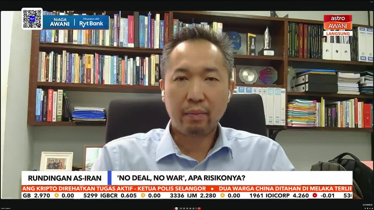 A man with earbuds speaks directly to the camera, framed by bookshelves. A news ticker scrolls across the bottom of the screen, displaying financial data and headlines in Malay.