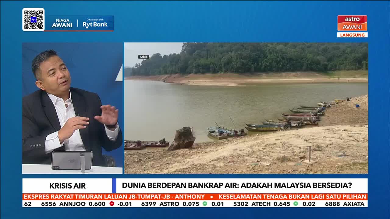 The water level in this Malaysian reservoir has dropped significantly, leaving many small boats resting on the exposed, cracked earth. A man in a suit gestures while speaking on Astro Awani, discussing the water crisis.