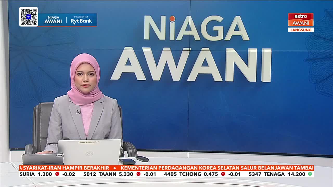 A woman in a pink hijab and grey blazer sits at a desk, looking directly ahead. Behind her, large white letters spell out "NIAGA AWANI" against a blue background.