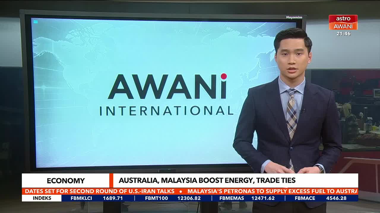 A young man in a suit stands before a large screen displaying "AWANI INTERNATIONAL." Below him, a ticker scrolls with news headlines, mentioning "AUSTRALIA, MALAYSIA BOOST ENERGY, TRADE TIES."